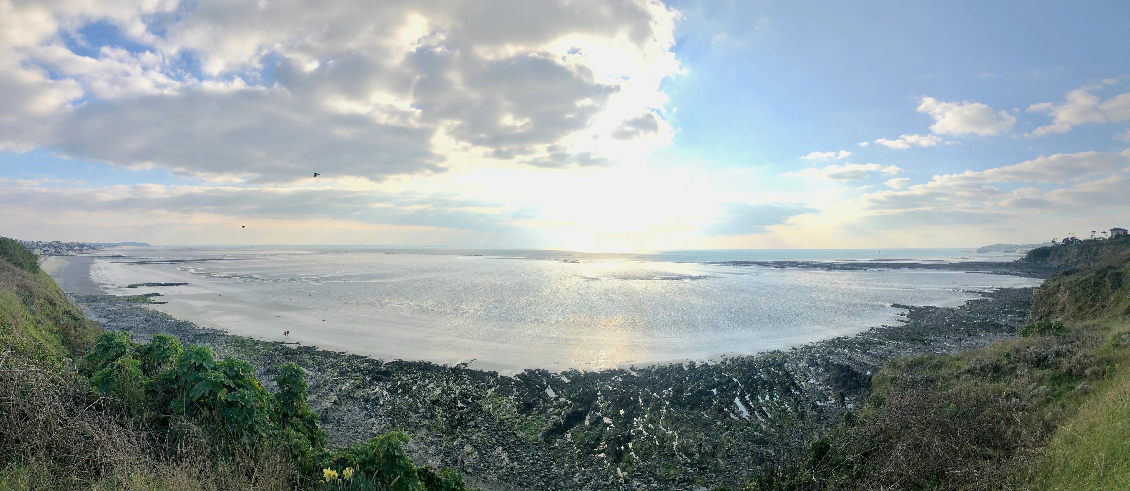 La baie à marée basse vue du haut de la falaise : plage de sable, rochers découverts et soleil bas sur la mer